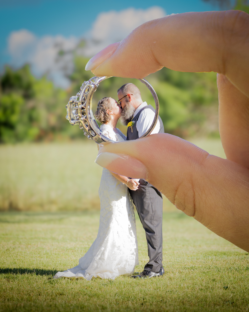 rustic-outdoor-wedding-texas-bride-groom-kiss A bride and groom kissing in a field as seen through a wedding band.